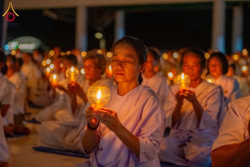 อุบาสิกาแก้วสวมชุดสีขาวนั่งหลับตาถือประทีปเทียนในมือท่ามกลางกลุ่มผู้เข้าร่วมพิธี