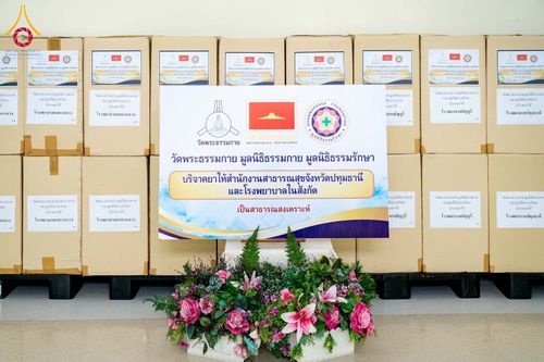 กล่องบรรจุยาจำนวนมากวางเรียงซ้อนกันพร้อมป้ายโครงการบริจาคยาเพื่อสาธารณสงเคราะห์โดยวัดพระธรรมกายและมูลนิธิที่เกี่ยวข้อง