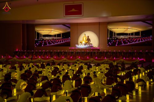 กลุ่มคนในชุดขาวนั่งปฏิบัติธรรมท่ามกลางแสงเทียนภายในห้องโถงกว้างที่มีจอภาพแสดงภาพการจุดเทียน