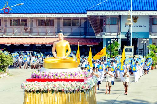 พระพุทธรูปสีทองบนฐานดอกไม้ในขบวนแห่ท่ามกลางกลุ่มคนที่เดินตาม