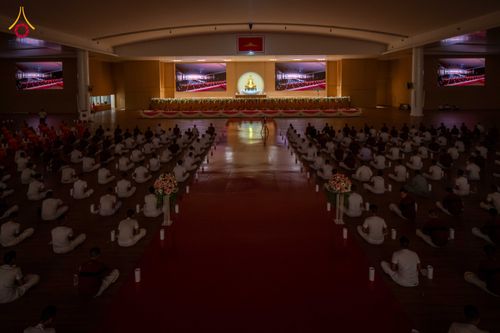 กลุ่มคนสวมชุดสีขาวนั่งสมาธิในห้องโถงกว้างขวางเพื่อร่วมพิธีจุดเทียนรวมใจ