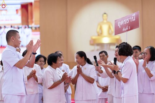 กลุ่มคนสวมชุดสีขาวกำลังยืนพนมมือและถือไมโครโฟนในงานสัมมนา โดยมีป้ายชื่อจังหวัดอุดรธานีและพระพุทธรูปเป็นฉากหลัง