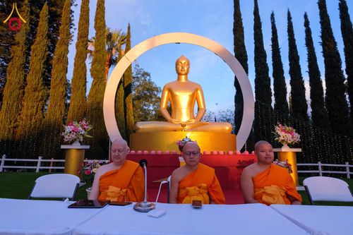 พระภิกษุสงฆ์สามรูปนั่งอยู่หน้าพระพุทธรูปสีทองขนาดใหญ่ในพื้นที่กลางแจ้ง