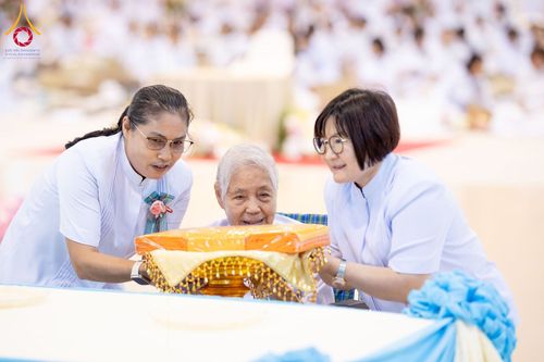 หญิงสูงวัยและเจ้าหน้าที่หญิงสองคนกำลังประคองพานผ้าไตรจีวรในพิธีทางศาสนา