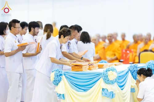 กลุ่มพุทธศาสนิกชนในชุดสีขาวกำลังวางผ้าไตรบนโต๊ะที่มีการตกแต่งด้วยผ้าสีฟ้าและสีเหลือง ในพิธีทางศาสนา