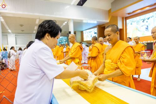 พระภิกษุสงฆ์ในจีวรสีเหลืองทองกำลังรับมอบพระของขวัญจากพุทธศาสนิกชนในชุดสีขาวภายในอาคาร