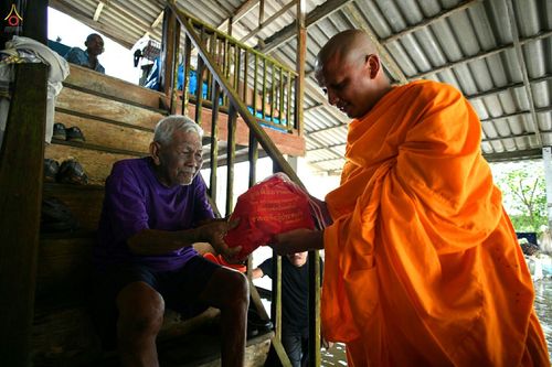 พระสงฆ์กำลังส่งมอบถุงยังชีพสีแดงให้กับชายสูงอายุที่นั่งอยู่บนบันไดไม้ของบ้านท่ามกลางสถานการณ์น้ำท่วม