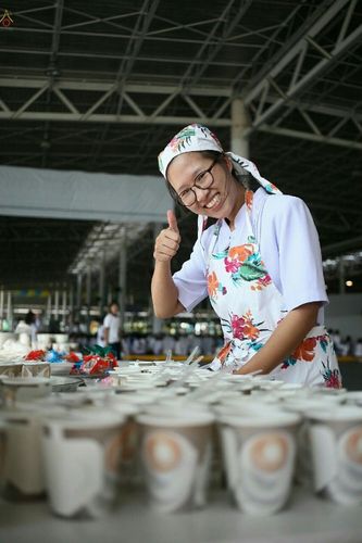 อาสาสมัครหญิงสวมผ้ากันเปื้อนลายดอกไม้กำลังเตรียมถ้วยอาหารและชูนิ้วโป้งด้วยรอยยิ้มในกิจกรรมบุญวันอาทิตย์ต้นเดือน