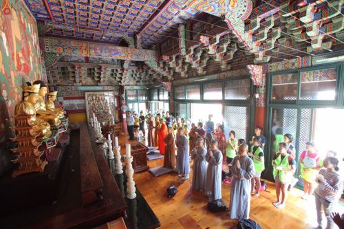 กลุ่มเยาวชนและพระสงฆ์กำลังยืนพนมมือสวดมนต์หน้าพระพุทธรูปภายในวัดพุทธนิกายโจเกที่มีการตกแต่งเพดานและผนังด้วยลวดลายศิลปะแบบดั้งเดิม