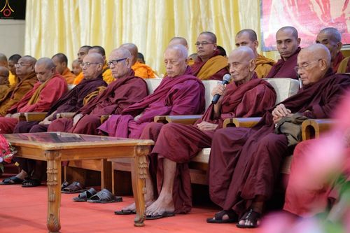 คณะสงฆ์นั่งเรียงแถวบนเก้าอี้ในพิธีทางศาสนาพุทธ โดยมีพระเถระถือไมโครโฟนกล่าวในงานพิธีถวายเทียนพรรษาและมหาสังฆทาน 111 วัด