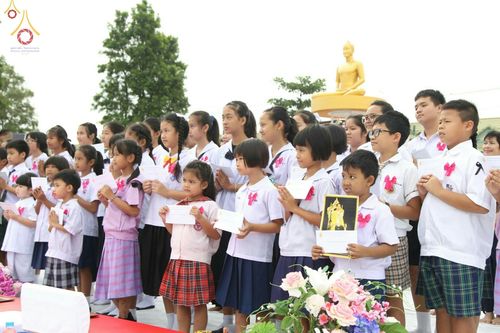 กลุ่มนักเรียนยืนพนมมือถือแผ่นกระดาษและรูปภาพในพิธีตักบาตรพระสงฆ์