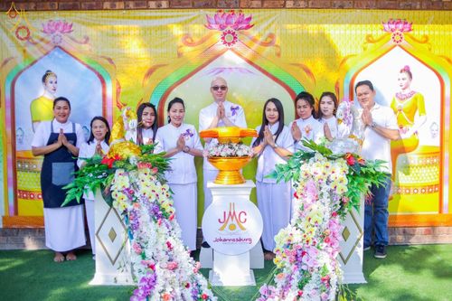 กลุ่มสาธุชนยืนพนมมือถ่ายภาพร่วมกันหน้าพานพุ่มดอกไม้และป้ายวัดพระธรรมกายโจฮันเนสเบิร์กในพิธีฉลองอุโบสถและทอดกฐิน