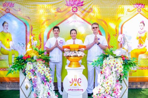 พุทธศาสนิกชนชายหญิงในชุดขาวร่วมพิธีทางศาสนาพุทธหน้าพานพุ่มดอกไม้และฉากหลังสีเหลืองทอง ณ วัดพระธรรมกายโจฮันเนสเบิร์ก