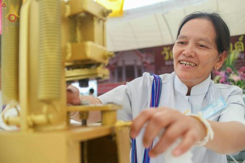 หญิงไทยสวมชุดขาวกำลังใช้เครื่องกดพิมพ์พระของขวัญด้วยความตั้งใจและมีรอยยิ้ม ในพิธีพิมพ์พระของขวัญรุ่นบรรลุธรรม