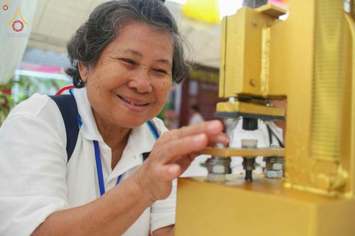 หญิงสูงวัยกำลังใช้เครื่องมือพิมพ์พระของขวัญรุ่นบรรลุธรรมด้วยความตั้งใจและมีรอยยิ้ม