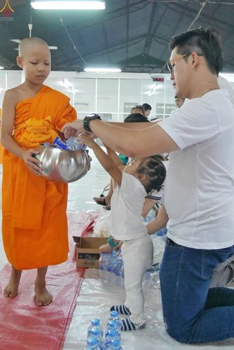 ชายและเด็กเล็กกำลังใส่สิ่งของลงในบาตรของสามเณรใหม่ภายในอาคารอบรม