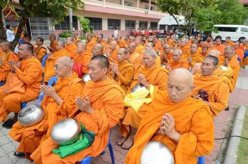 พระภิกษุสงฆ์จำนวนมากในจีวรสีส้มกำลังนั่งพนมมือสำรวมในพิธีตักบาตรที่โรงเรียนเบญจมราชูทิศ จังหวัดปัตตานี