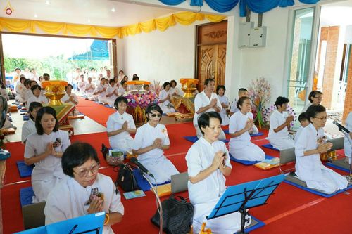 กลุ่มพุทธศาสนิกชนในชุดขาวนั่งพนมมือประกอบพิธีทางศาสนาภายในอาคารที่มีการตกแต่งด้วยผ้าสีเหลืองและสีน้ำเงิน