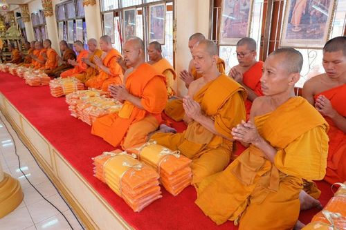 คณะสงฆ์นั่งพนมมือประกอบพิธีทางศาสนาและชุดผ้าไตรที่วางเรียงรายบนพรมสีแดง