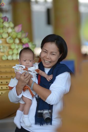หญิงไทยยิ้มแย้มอุ้มทารกน้อยในอ้อมแขนภายในงานบุญที่มีการประดับตกแต่งด้วยดอกบัว