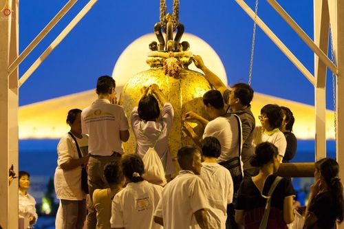 พุทธศาสนิกชนกำลังปิดแผ่นทองคำเปลวบนระฆังธรรมขนาดใหญ่ท่ามกลางบรรยากาศยามเย็น