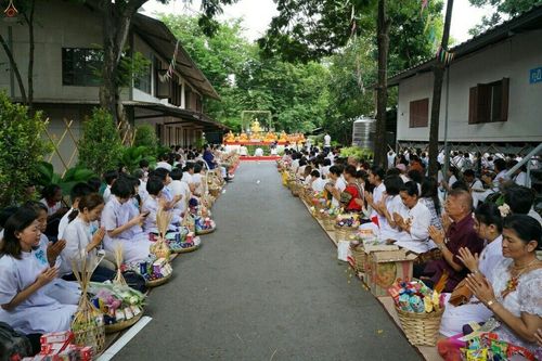 พุทธศาสนิกชนสวมชุดขาวนั่งเรียงรายสองฝั่งถนนเตรียมตักบาตรพระสงฆ์ในพิธีตักบาตรย้อนวันวาน