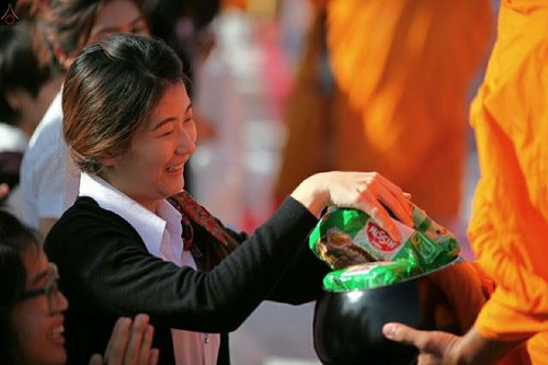 หญิงสาวกำลังใส่บะหมี่กึ่งสำเร็จรูปในบาตรพระภิกษุสงฆ์ด้วยใบหน้ายิ้มแย้มในงานตักบาตรกตัญญู