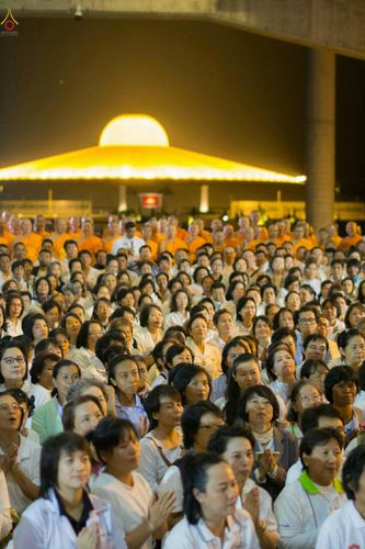 พุทธศาสนิกชนจำนวนมากสวมชุดขาวร่วมประกอบพิธีสวดธัมมจักกัปปวัตนสูตร โดยมีพระภิกษุสงฆ์ร่วมในพิธี ท่ามกลางบรรยากาศยามค่ำคืนที่มีฉากหลังเป็นมหาธรรมกายเจดีย์ส่องสว่าง
