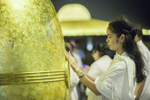หญิงสาวสวมชุดขาวกำลังปิดทองระฆังธรรมสีทองขนาดใหญ่ในพิธีทางศาสนา