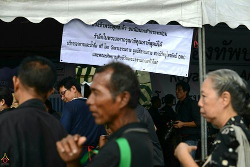 ประชาชนเดินผ่านจุดบริการอาหารและน้ำดื่มฟรีที่มีป้ายข้อความรำลึกในพระมหากรุณาธิคุณ