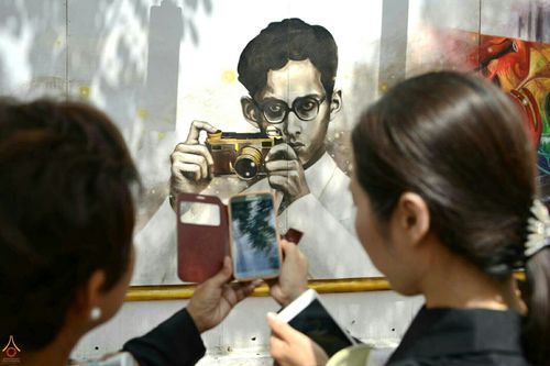 ประชาชนกำลังใช้โทรศัพท์มือถือถ่ายภาพจิตรกรรมพระบรมสาทิสลักษณ์ของในหลวงรัชกาลที่ 9 ขณะทรงกล้องถ่ายภาพ