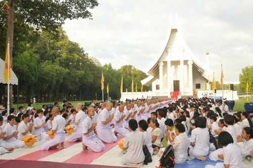 กลุ่มพุทธศาสนิกชนและนาคในชุดขาวนั่งพนมมือร่วมพิธีบรรพชาอุปสมบทหมู่นาคธรรมทายาทหน้าอาคารทรงไทย