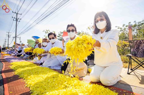 พุทธศาสนิกชนสวมชุดขาวนั่งเรียงแถวโปรยกลีบดอกดาวเรืองสีเหลืองต้อนรับพระธรรมยาตรา