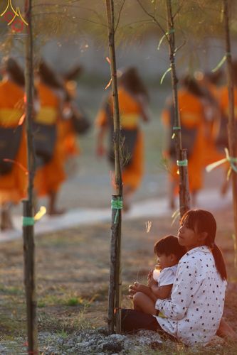 หญิงสาวอุ้มเด็กนั่งรอชมขบวนพระธรรมยาตราที่กำลังเดินผ่านในพื้นที่โล่งแจ้งยามเย็น