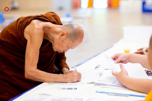 พระภิกษุสงฆ์กำลังเขียนเอกสารลงทะเบียนสอบธรรมสนามหลวงบนโต๊ะที่มีเจ้าหน้าที่คอยดูแล