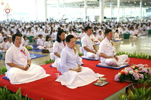 กลุ่มพุทธศาสนิกชนสวมชุดขาวนั่งสมาธิปฏิบัติธรรมบนอาสนะสีแดงภายในอาคารขนาดใหญ่