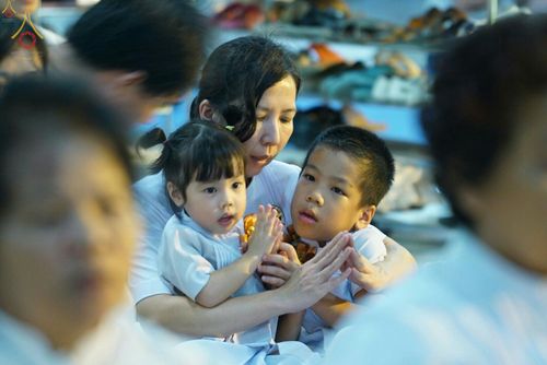 หญิงชาวไทยสวมชุดขาวโอบกอดลูกชายและลูกสาวที่กำลังพนมมือไหว้ในกิจกรรมทางศาสนา